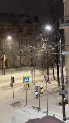 The video depicts a winter scene at night in what appears to be a small town or city in Russia, with snow falling steadily. A street intersection is visible, covered in snow, illuminated by streetlights and traffic lights. Several road signs are present, including a pedestrian crossing sign, a yield sign, and a stop sign written in Russian. A yellow taxi drives down the street and turns at the intersection. Buildings line the street, including one with a balcony, and bare trees are visible against the night sky. The overall atmosphere is quiet and snowy.