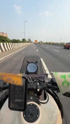 This media file captures a first-person perspective of a motorcycle ride on a highway, likely in Samalkha, India. The rider is wearing a red helmet with a black visor and a black and red riding jacket. They are also wearing black gloves and appear to be giving a peace sign with their left hand. The motorcycle has a digital display showing "67" which could be the speed in km/h. Prayer flags, one yellow with the mantra "Om Hum" and another blue with "Om," are attached to the handlebars.

The scene is a multi-lane highway with concrete barriers and streetlights. The weather appears clear and sunny, suggesting it is daytime, likely late afternoon given the angle of the sun. Other vehicles, including trucks and cars, are visible on the highway, indicating active traffic. Trees and some buildings are visible in the background, bordering the highway. The overall mood conveyed is one of travel and exploration. The text visible includes "CHOKKAS" on a black box on the motorcycle and "AERONAUTICS" on the rider's helmet.