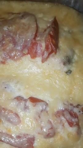 This is a close-up, static shot of a dish containing tomatoes and melted cheese. The dish appears to be a baked or gratin dish, with the cheese melted and bubbly over the tomatoes. The lighting suggests it might be indoors, possibly in a kitchen or dining area. The image is a bit grainy and the focus is not perfectly sharp, but the textures of the melted cheese and the softened tomatoes are clearly visible. There is no visible text, and no people are present in the frame. The exact time of day and weather conditions are not discernible from the image. Given the provided location context of Shklov, Belarus, it can be inferred this is likely a homemade meal or a dish served in a local eatery. The overall impression is one of comfort food.