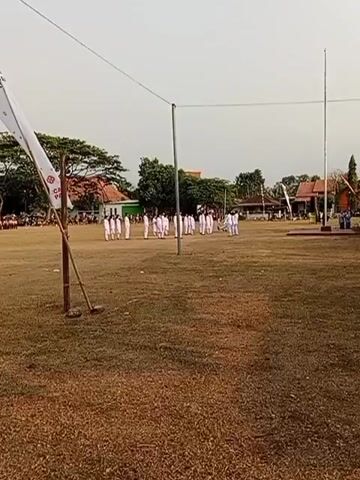 The video shows an outdoor scene on a dry, brown field. In the foreground, a white flag with red text, partially obscured by a wooden pole, flutters to the left. In the middle ground, a group of individuals, dressed in white, stand in formation on the field. A tall, thin pole stands in the center of the frame, with a taut wire extending from it to the left. In the background, several buildings with red roofs are visible, along with trees and a clear sky. To the right, another tall pole, presumably a flagpole, is present. The overall impression is that of an outdoor event or gathering, possibly a school ceremony or assembly.