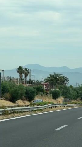 A car is driving on a multi-lane highway under an overcast sky. The highway has a wide median with vegetation and guardrails. In the distance, there are gently sloping mountains. Palm trees and other greenery are visible on the side of the highway. Overhead, there's a bridge with a pedestrian walkway. Cars are driving in both directions. Road signs, including a blue arrow pointing straight ahead and a red "do not enter" sign, are placed on the sides of the road.