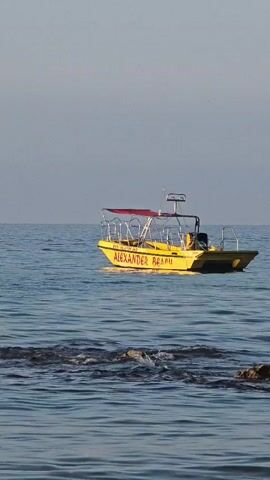 A yellow boat with a red canopy is floating on the sea. The boat has the words "ALEXANDER BEACH" written on its side. The water is calm and the sky is clear. In the distance, another boat is visible with a few people in it. There is also a small structure, possibly a gazebo, built on rocks in the water. The overall scene is peaceful and serene, suggesting a calm day at sea.