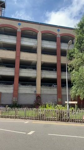 The video shows a multi-story parking garage with a red and beige facade, featuring arched openings on each level. A light pole stands in front of the garage, partially obscured by trees. A wooden fence with a white picket design runs along the front of the property, separating it from a paved road. Behind the fence, there's a small garden area with various plants and some decorative elements, including a small stone structure. The trees vary in size and species, with one being particularly tall and dense, casting shadows on the garage. The sky above is partly cloudy with a blue hue. The camera pans slightly to the right, revealing more of the garage and the surrounding trees. No people or vehicles are clearly visible or engaged in any action. The scene appears to be an exterior shot on a daytime, likely sunny, day.