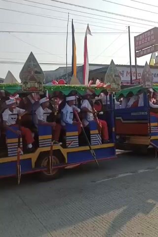 The video shows a parade of colorful, open-air vehicles resembling modified jeeps. The vehicles are decorated with flags, balloons, and what appear to be traditional puppet figures at the front. Several people, mostly young males dressed in white shirts and orange pants, are seated in the vehicles, some holding long poles or oars. The vehicles are driving on a paved road, with buildings and power lines visible in the background. Two motorcycles are also present in the foreground, one carrying two people. The overall atmosphere suggests a festive or celebratory event.