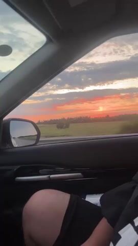The video shows a person's legs and arm resting inside a car, with a view out the window. The scene outside the window is a rural landscape with a sunset featuring orange and pink clouds. The car appears to be in motion, as indicated by the blurred background and the changing perspective of the rearview mirror. The interior of the car includes a dashboard and a seatbelt.