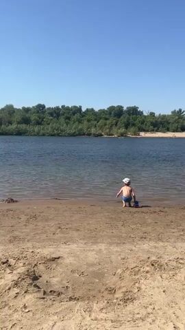 A young child wearing a white hat and blue swim trunks is crouched on a sandy beach near the edge of a wide, calm river under a clear blue sky. Dense green trees line the opposite bank of the river. A jet ski speeds across the water in the distance, creating a large spray of water behind it. The jet ski is moving from left to right across the frame.