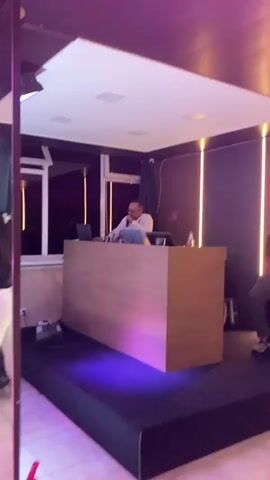 The video shows a dimly lit room with colored lights. A man wearing a white shirt and sunglasses stands behind a DJ booth, holding a microphone and appearing to speak. In the background, there are windows with vertical lights visible outside. The DJ booth is on a raised platform covered in black fabric, with green and purple lights projected onto it. Further into the room, several people are dancing, including a woman in a white, sparkly dress and a child with braided hair being held by a man. The overall atmosphere suggests a party or event.