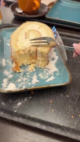 A fork is being used to cut into a slice of Swiss roll cake that has been placed on a teal-colored plate. The cake appears to be filled with a white cream and dusted with powdered sugar, some of which has spilled onto the plate. In the background, an orange coffee cup sits on a saucer, and another teal plate is visible. The setting appears to be a table, possibly in a cafe or restaurant.