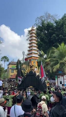 A religious procession is taking place on a sunny day, possibly in Southeast Asia, given the palm trees and traditional attire. A tall, multi-tiered tower, adorned with white, gold, and red decorations, is being carried by a group of people on a platform supported by poles. The tower has a distinctive pagoda-like structure. Below the tower, a golden, stylized sun-like decoration is visible, and the platform is partially covered by a black, arched structure. Numerous people, many wearing green shirts and traditional sarongs, are surrounding the platform, some holding onto the poles to carry the structure. Others are standing in the background, observing the procession. Flags, including one red and white flag, are also part of the display. The environment appears to be a street lined with buildings and lush green trees, suggesting an urban or semi-urban setting. The sky is blue with scattered white clouds. Some individuals in the crowd are wearing face masks. The overall atmosphere suggests a festive or ceremonial event.