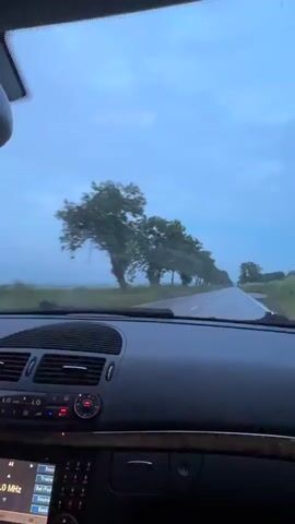 This video shows the view from inside a car driving on a road with trees lining both sides. The sky is overcast and appears to be raining, as evidenced by the blurred windshield wipers and the misty atmosphere. The dashboard of the car is visible in the foreground, with some buttons and a screen displaying information. The scenery outside is green with vegetation.