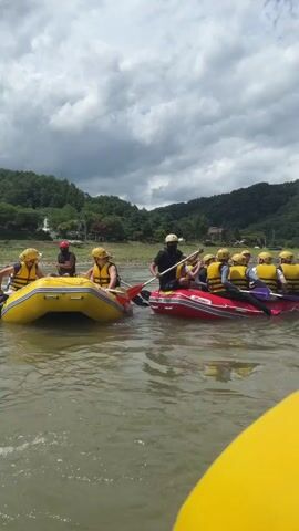 A group of people are white-water rafting on a river. The camera is in a yellow raft, and in the foreground, we see the yellow inflatable sides of the raft and a person's hands holding a paddle. In the middle ground, another raft with several people wearing yellow helmets and life vests is visible. One person in that raft, wearing a blue and black patterned shirt and a yellow life vest, is taking a picture with a phone. The background consists of a green, tree-covered hillside under a cloudy sky. The water is calm.