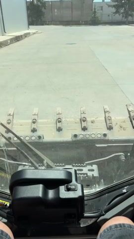 This is a first-person perspective video from inside a vehicle, likely a skid steer loader, given the visible controls and the front attachment. The operator's legs, clad in denim shorts, are visible at the bottom of the frame.

The **scene** is an outdoor area with a paved surface, possibly a factory yard or industrial site. In the background, there are buildings with brick walls and some trees. The sky is blue, suggesting it is daytime with clear weather. The overall atmosphere is that of an operational environment.

The **activity** appears to be the movement or operation of the vehicle. The camera is focused on the front attachment, which looks like a bucket or a grapple with several teeth. The windshield of the vehicle is visible, along with the wiper mechanism. A black control lever with buttons is prominent in the foreground, likely for operating the vehicle's functions.

**Notable details** include the dusty condition of the front attachment, indicating recent use. The bright sunlight and clear sky suggest a warm, fair-weather day. The buildings in the background with their industrial architecture provide context about the location.

There is **no visible text** in the media file. The time of day is clearly daytime due to the bright sunlight and clear blue sky. The location cues (industrial buildings, paved yard) and the type of vehicle suggest an industrial or construction setting. No specific emotions are conveyed, as the focus is on the operational aspect.