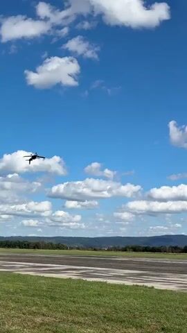 This is a vertical, point-of-view video capturing a clear blue sky with scattered white, fluffy clouds. A small, dark-colored airplane is seen flying horizontally in the upper portion of the frame. Below the sky, a flat, open landscape is visible, featuring a tarmac or runway area with green grass bordering it. In the distance, there are rolling hills or mountains under the horizon. The airplane appears to be in mid-flight, with no visible interaction with any people or ground objects. The overall impression is of an aerial view of a model airplane, possibly at an airfield.