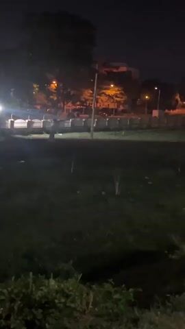 The video shows a dark, outdoor scene at night, possibly a park or grassy area. In the background, there are blurred lights from street lamps and buildings. At one point, a bright light shines directly at the camera, causing a lens flare. A person wearing a white shirt briefly appears in the foreground. The overall setting appears calm and quiet.