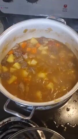A pot of beef stew is simmering on a stovetop. The stew contains chunks of beef, potatoes, and carrots in a rich brown broth. Steam is rising from the pot, indicating it is hot and cooking. A green spatula is visible stirring the stew. The stovetop is black and appears to be electric, with control knobs visible at the top.
