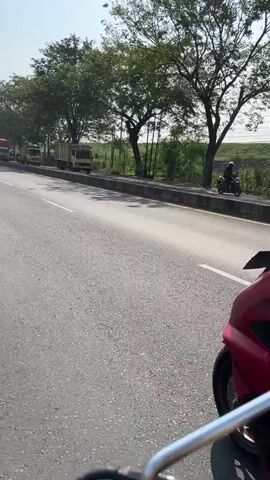 A person wearing a helmet and a light blue shirt rides a red motorcycle. They have a large black backpack strapped to their back, and a long, thin red case is attached to the top of the backpack, extending upwards. The motorcycle has a license plate that reads "S 3958 NCF". The motorcycle is on a paved road with a white line marking the lanes. In the background, there are trees, a bright sky, and several vehicles, including a red truck. The sun is shining, casting shadows.