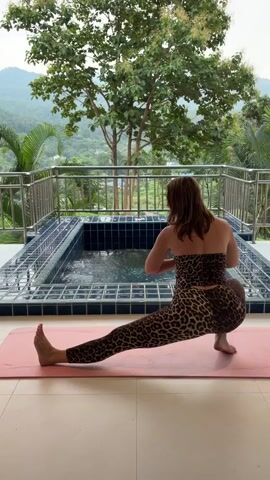A woman in leopard print athleisure wear performs a side lunge yoga pose on a pink yoga mat. She is positioned on a balcony with a view of a lush, green landscape and mountains in the distance. A small, rectangular swimming pool with blue tiles is visible behind her. The woman's right leg is extended to the side with her foot flat on the mat, while her left leg is bent with her knee close to the floor. Her hands are clasped in front of her chest. The sun is out, casting a soft light on the scene.