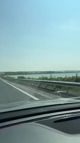 The video shows a first-person perspective from inside a car driving on a highway. The road is asphalt with white lane markings. To the right of the road, there is a metal guardrail and then a body of water with trees and vegetation along its edge. The sky is clear and blue. The dashboard of the car is visible in the foreground. The car appears to be moving at a moderate speed.