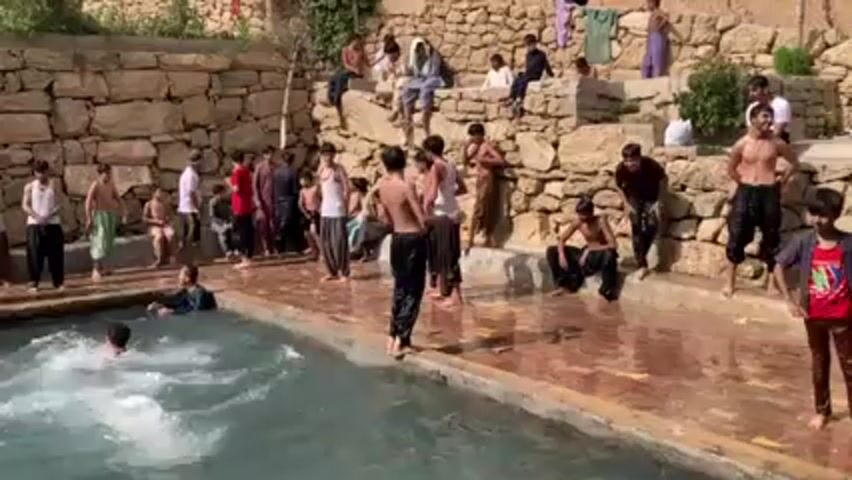 In an outdoor setting, a group of young males enjoys a swim in a rectangular body of water, possibly a pool or a natural spring. The water's color is light blue. Most of the young men stand around the pool's edge, some shirtless and wearing dark pants, while others are fully dressed. A few are already swimming, creating splashes in the water.  

Behind the group, there's a high, stone-wall structure with more people, including adults and children, sitting and observing the swimmers. The wall suggests a terraced or mountainous landscape. The overall atmosphere seems cheerful, with the young men actively engaging in water activities under the open sky.