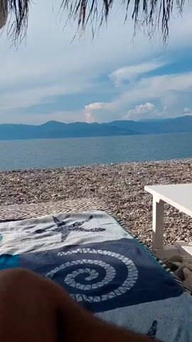 The video shows a first-person view of someone lying on a beach chair under a thatched umbrella. In the foreground, a patterned towel with a starfish and a spiral design is draped over the chair. To the right, a corner of a white table is visible. Beyond the chair and table, a pebble beach extends to the edge of a calm, blue sea. In the distance, a range of hazy mountains forms the horizon under a sky with scattered white clouds. The overall impression is one of a relaxed beach scene.