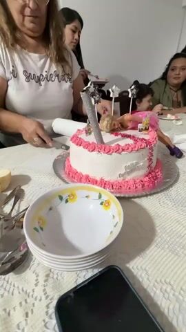A group of people are gathered around a table on which sits a Barbie-themed birthday cake. A woman wearing a white shirt with writing is using a knife to cut into the cake, which is decorated with pink frosting and has a Barbie doll lying on top. There are bowls and silverware on the table, and a smartphone is visible in the foreground. Other individuals are seated around the table, with a child wearing a red shirt visible among them. The atmosphere appears to be celebratory, likely a birthday party.