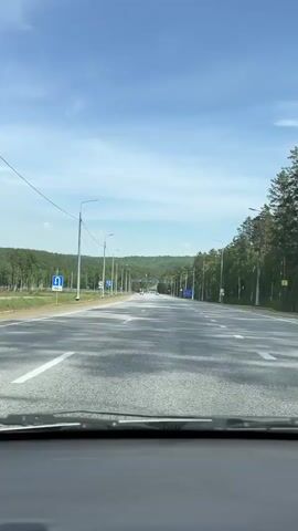 The video is a first-person perspective from inside a vehicle, looking out through the windshield. The vehicle is driving on a paved, multi-lane highway that appears to be flanked by a forest of pine trees on the right and a more open area with some sparse vegetation and infrastructure on the left. The sky is blue with scattered white clouds. There are traffic signs visible on both sides of the road, including blue directional signs and speed limit signs. Streetlights line the sides of the highway. The road has clear white lane markings. In the distance, there is a slight hill or mountain range covered in trees. The sun is casting shadows on the road from the trees.