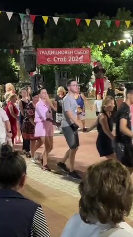 The video captures a lively outdoor event with a group of people dancing in a circle. A banner in the background reads "ТРАДИЦИОНЕН СЪБОР с. Стоб 2024," indicating a traditional gathering in the village of Stob in 2024. The dancers, a mix of men and women, are holding hands and moving rhythmically. They are dressed in casual and festive attire. A musician can be seen on a platform in the background, possibly playing traditional music for the dance. The setting appears to be a town square or a public gathering space, decorated with colorful triangular flags. The overall atmosphere is one of celebration and community engagement. The camera is positioned behind some spectators, providing a glimpse of the event from their perspective.