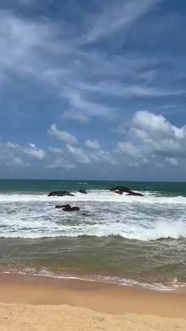 This is a vertical video of a beach. The foreground shows a sandy shore. The middle ground features the ocean with waves breaking on dark rocks. The background displays a blue sky with white clouds.