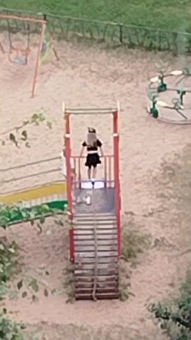 A young girl in a black dress stands at the top of a playground slide. Another child, a boy in a white shirt, approaches her from the stairs leading up to the slide. The girl turns to face him, and they appear to interact briefly. The video is filmed from an elevated perspective, showing a sand-covered playground with a swing set and a merry-go-round in the background. Trees and greenery are visible on the left side of the frame.