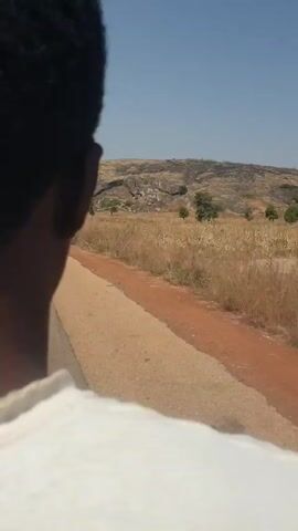 The video depicts a person driving or riding along a road with dry, grassy fields on the right and a rocky hill in the background under a clear blue sky.  The road has both paved and dirt sections. The field appears to be overgrown with dry grass and some small trees.
