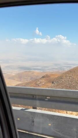 A video shot from inside a vehicle shows a view through the window. The foreground features a metal guardrail. Beyond the guardrail, a dry, mountainous landscape stretches out. In the distance, a hazy plain is visible under a blue sky with scattered white clouds. The overall impression is of a scenic drive through a semi-arid region.
