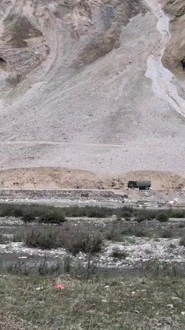 A truck drives along a dirt road that cuts through a barren, mountainous landscape. The truck is dark-colored and appears to be military or industrial in nature. It is moving from left to right across the frame. There are two smaller white vehicles parked on the side of the road to the left of the truck. The area is dry and rocky, with sparse vegetation. A small stream or riverbed runs through the foreground. In the distance, the slopes of the mountains are covered in scree and loose rock. The sky is overcast and the lighting suggests it is daytime.