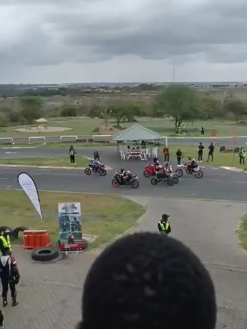 The video captures a motorcycle race taking place on a track, viewed from an elevated position. Several motorcycles are seen speeding along the track, accompanied by a pace car. The track is bordered by green grass and trees, with a cityscape visible in the background under a cloudy sky. The environment also includes signs and structures, such as a tent with people seated underneath it. The video transitions to show the motorcycles speeding further away.