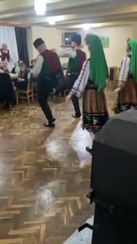 A video shows a group of people in traditional Bulgarian folk costumes performing a line dance in a well-lit indoor space with a wooden parquet floor. The dancers, including two men with small black hats and green vests embroidered with red, move in unison. Women dancers, wearing white tops with embroidered sleeves, black skirts with colorful patterns, and green head coverings, are also visible. The setting appears to be a gathering or celebration, with tables and chairs occupied by seated individuals in the background. A large black speaker box is in the foreground, slightly obscuring the view of the dance. The video is taken at a close-range and follows the movement of the dancers.