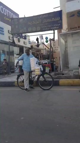 The video pans from left to right showing a street scene in what appears to be a Middle Eastern town.  The first store visible has a sign that says "Cottonil" in English and Arabic.  The next store is called "New Smile" and also advertises cotton goods.  The third store has signage in Arabic and appears to sell plumbing and sanitary supplies.  People are seen walking on the sidewalk and crossing the street.  The sky is visible at the top of the frame.