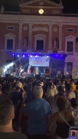 The video shows an outdoor concert at night. A band named "Tarraco Surfers" is performing on a stage in front of a large crowd of people. The stage is lit with blue and purple lights. The band members are visible on stage, playing instruments and singing. The crowd is densely packed, and many people are looking towards the stage. Behind the stage, there is a large building with arched windows and a clock on its facade. The overall atmosphere appears to be lively and energetic.