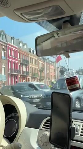 The video, filmed from inside a car, captures a street scene during daylight. The interior of the car is visible, including the dashboard, rearview mirror, and a smartphone mounted on a stand. Hanging from the rearview mirror is a small jar with a red substance, possibly an air freshener. Through the windshield and side windows, a street with buildings, other cars, and a street lamp can be seen. The sky is blue with some clouds. The video appears to be a continuous shot, possibly from a phone mounted on the dashboard.