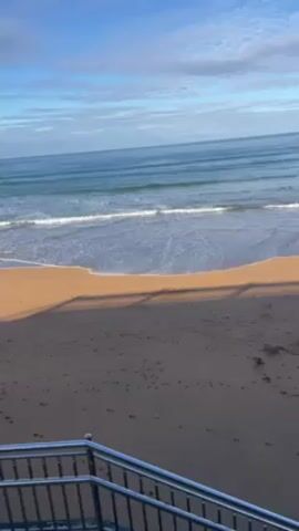 The video shows a beach on a sunny day. The sea is calm with small waves. A set of metal railings is in the foreground, casting a shadow on the sand. The sand is brown and has footprints on it. The sky is blue with some clouds. The camera pans slightly.
