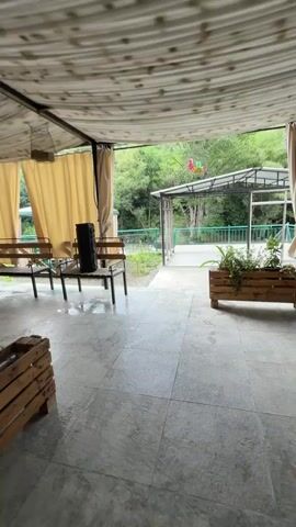 This media file captures a peaceful outdoor scene, likely a seating area at a restaurant or cafe. The foreground shows a covered patio with a tiled floor, appearing slightly damp, suggesting recent rain or cleaning. A black speaker sits on a wooden table next to two chairs, hinting at the possibility of music or entertainment.

Beyond the patio, the scene opens up to a lush green landscape. Trees and foliage cover rolling hills, creating a backdrop of natural beauty. A river or stream flows in the middle ground, adding to the serene atmosphere. Several gazebos or covered seating areas are visible, some with tables and chairs, suggesting a place for patrons to relax and enjoy the surroundings. A metal railing lines the edge of the seating areas, separating them from the natural environment. In the distance, a small structure with a white roof and metal supports stands. A colorful object, possibly a kite or decoration, is suspended in the air above this structure.

The overall impression is one of tranquility and relaxation. The cloudy sky suggests it might be overcast or late afternoon. There is no visible text in the media file. The location context of Alaverdi, Armenia, further evokes a sense of being in a natural, possibly mountainous, region.