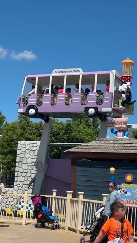 The video shows a purple bus ride at an amusement park. The bus, labeled "El autobús de Piolín," is filled with people and elevated on an arm, rotating in a circular motion. On top of a nearby building, there is a statue of Porky Pig holding a cake. Below it, there is a sign that reads "Piolín y Sylvester Paseo En Autobús." In the foreground, a woman is standing with two strollers near a white picket fence. A boy in an orange shirt is visible to the right. The sky is clear and blue.