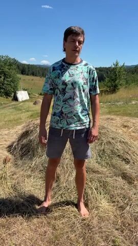 A man with short brown hair stands on a haystack with his arms outstretched in a rural, outdoor setting. The sky is blue and clear. The man is wearing a teal and white patterned t-shirt and grey shorts. He is barefoot. Behind him, the landscape consists of rolling hills, trees, and a small white building. The man then falls backwards onto the haystack, lying down with his arms still outstretched. He appears to be enjoying himself.