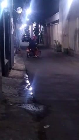 A person wearing a red helmet and a dark jacket rides a red scooter down a dark alley at night. The scooter is on the right side of the alley. The rider is leaning over the handlebars. There are buildings on both sides of the alley. There are streetlights in the distance. The ground is wet and reflective.