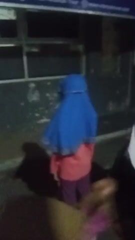 The video begins with a close-up of a young girl wearing a blue hijab and a pink shirt. She is looking away from the camera and her face is partially obscured. The background is dark and appears to be outdoors at night, with a building and windows visible. As the video progresses, the camera pans to the left, revealing other children sitting on the ground behind the girl. One boy is wearing a green shirt, and another person is lying down. The lighting is dim. The video then briefly shows a blurred image of someone walking away, followed by two girls in hijabs, one in white and the other in black and white, standing in an outdoor area. There are some chalk markings on the ground. The video concludes with a blurry shot of the girl in the blue hijab, who has now turned to face the camera, with other children visible in the background.