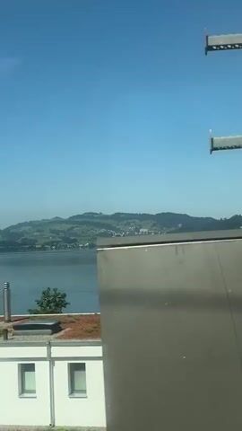 The video captures a scenic view of a calm lake under a clear blue sky. The camera pans slightly to the right, revealing a green, rolling hillside dotted with houses and trees on the far side of the lake. In the foreground, the top of a modern building with a green roof and solar panels is visible. The building has white walls and a gray rectangular vent. A section of a red-tiled roof with a chimney is also present. The overall impression is one of a peaceful, sunny day in a picturesque landscape.