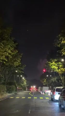 A car drives down a road at night, lined with trees and streetlights. In the distance, fireworks are being set off, visible as flashes of red light and smoke against the dark sky. The road is marked with yellow lines and there are traffic barriers and some parked cars on the side. The scene appears to be some sort of celebration or event.
