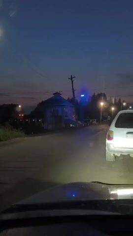 A view from inside a vehicle as it approaches a railway crossing at dusk. The sky is a deep blue with hints of orange near the horizon. A white car is stopped in front of the video-recording vehicle. Across the road, a red and white train is passing through the crossing. A red light flashes at the crossing signal. A small building with a dark roof is visible to the left of the crossing. Power lines and poles are present in the background. The road is asphalt and appears damp.