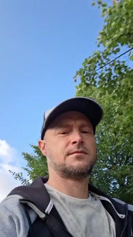 A medium close-up video begins with a man in a baseball cap and hoodie looking forward. He has short hair and stubble. Trees with green leaves are visible behind him, along with a blue sky. The camera then pans up towards the sky and to the right, revealing a streetlamp and some houses. The man's face is no longer fully visible in the frame, but he appears to be looking up. The video ends with the camera pointed at the sky.