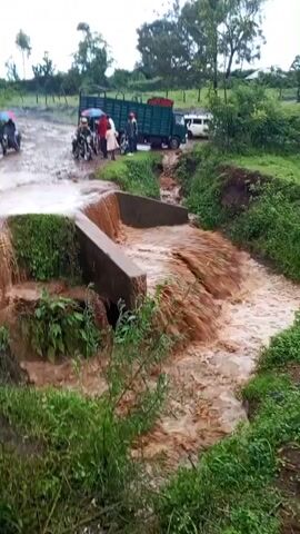 The video shows a flooded culvert with muddy water rushing through it. On the road, there is a truck, several people, motorcycles, and a tractor. The water overflows the culvert and is eroding the banks. The area appears rural with lush green vegetation. The sky is overcast. People seem to be observing the situation. The tractor begins to drive through the flooded road.