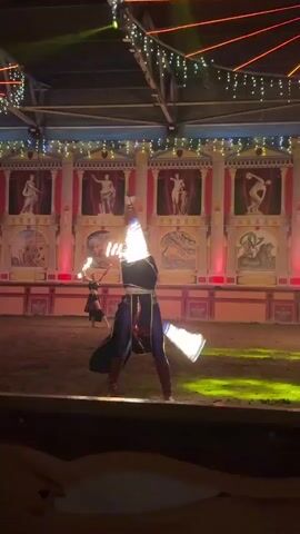 The media file captures a performance involving two individuals engaged in fire dancing. The scene is set in what appears to be an indoor arena with a sandy floor. In the background, a theatrical backdrop depicts classical Roman architecture with statues and murals, illuminated by red and yellow lights. The ceiling is adorned with festive string lights.

One performer, in the foreground, is dressed in a dark top with metallic accents and a long, flowing blue and red skirt, paired with red boots. This individual is actively twirling fire props, creating bright, fiery arcs in the air. To the left, another performer, wearing a black outfit with a skirt, is also seen manipulating fire props, leaving trails of light.

The lighting suggests it is evening or indoors, emphasizing the glow of the fire. The atmosphere seems energetic and captivating, as suggested by the dynamic movements of the performers and the visual spectacle of the fire. The overall impression is that of a dramatic and entertaining performance, possibly at a festival or a themed event. There is no visible text in the media file. The location context of Kamchatka, Russia, does not offer any specific visual cues within the provided frames.