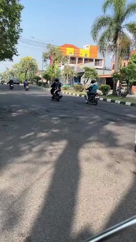 The video shows a street scene from the perspective of someone on a motorcycle. The road is paved and has some wear and tear. On the right side of the road, a white car is parked. In the middle ground, several motorcycles are visible, with riders wearing helmets. One rider in the foreground is wearing a black hoodie with a white star design. There are buildings on the left and right sides of the street, some of which are brightly colored. Trees and power lines are also present. The sky is clear and blue, suggesting daytime. The overall impression is a busy street in an urban or suburban area.